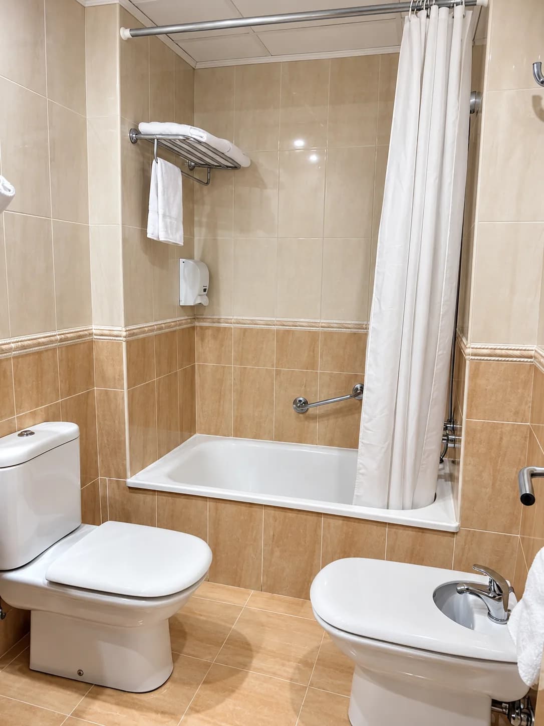 Baño privado con bañera, bidé y váter en azulejos beige del Hotel Plaza Inn en España, con toallas y cortina blanca cerrada
